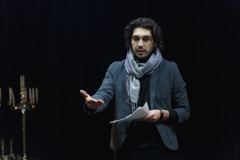Actor rehearsing on a dark theater stage, holding a script and gesturing dynamically.